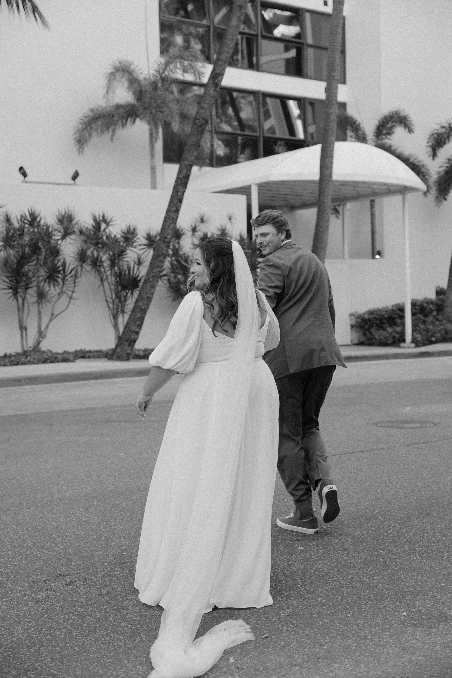 A playful couple in white dress and dark suit walk outdoors near palm trees.