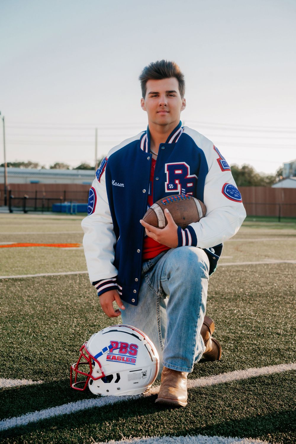 Athlete in varsity jacket kneeling with football and helmet on field.
