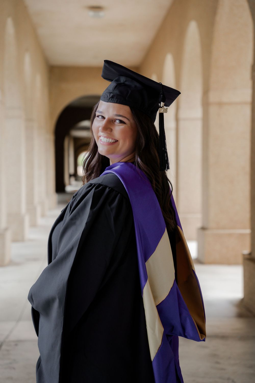 Graduate wearing black cap and gown with purple stole posing in archway.