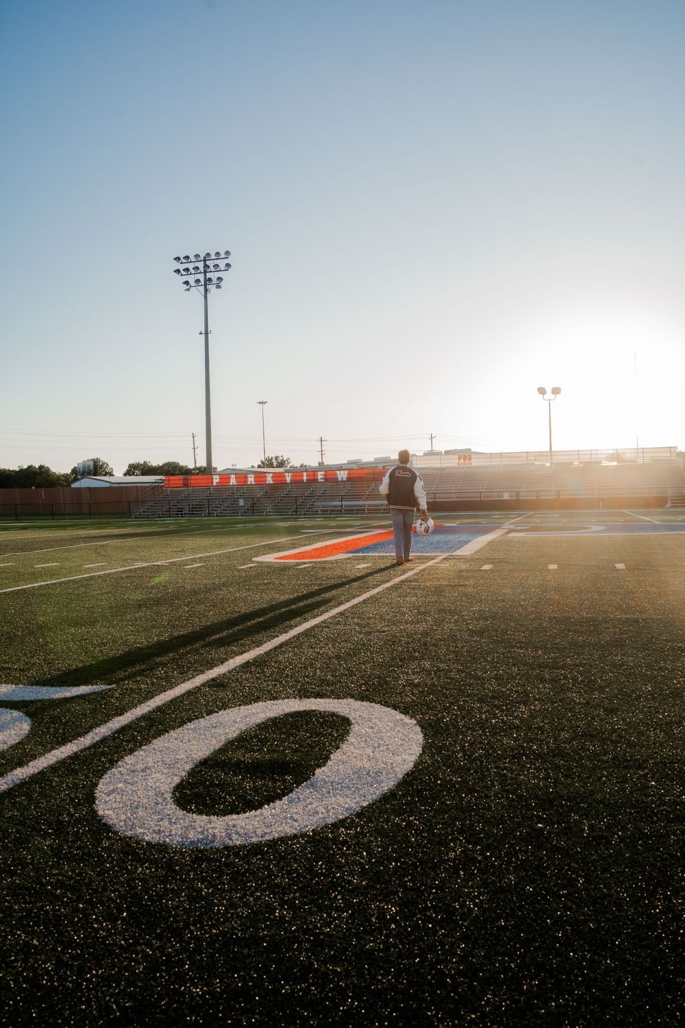 Football field at sunset with stadium lights and yard line markers visible.