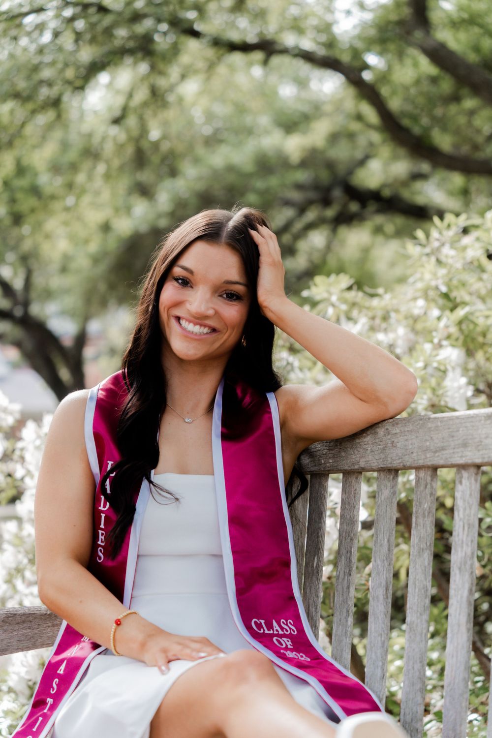 Graduate wearing pink and white graduation stole sitting on wooden bench surrounded by flowering trees.