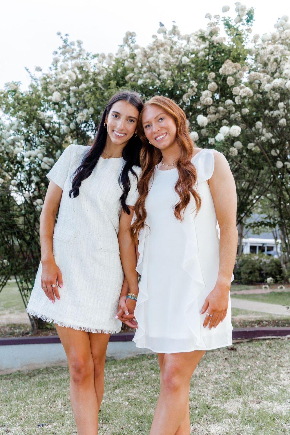 Two graduates in white dresses pose together in front of flowering bushes.
