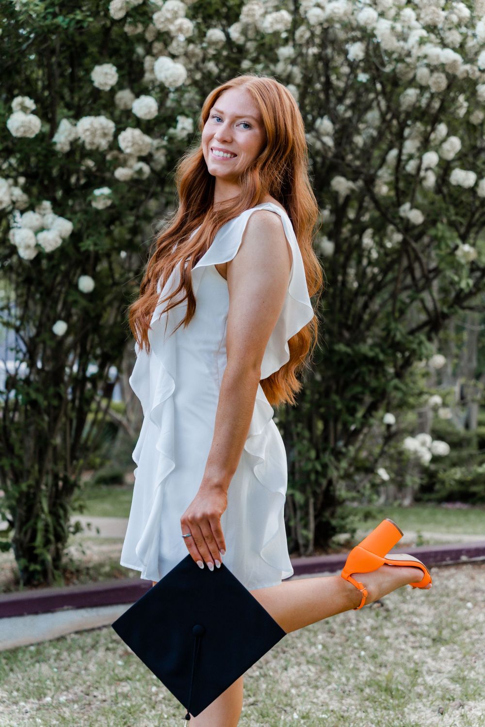 Graduate in white dress holds diploma while posing by blooming white roses.