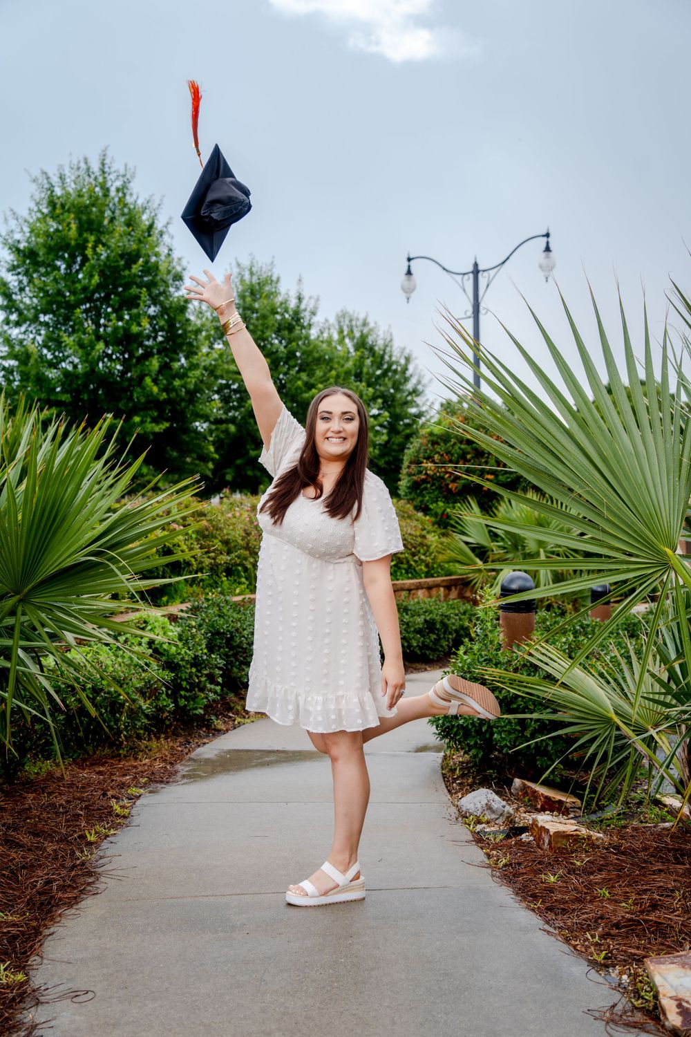 Graduate in white dress tossing graduation cap in the air on garden path with palm plants.