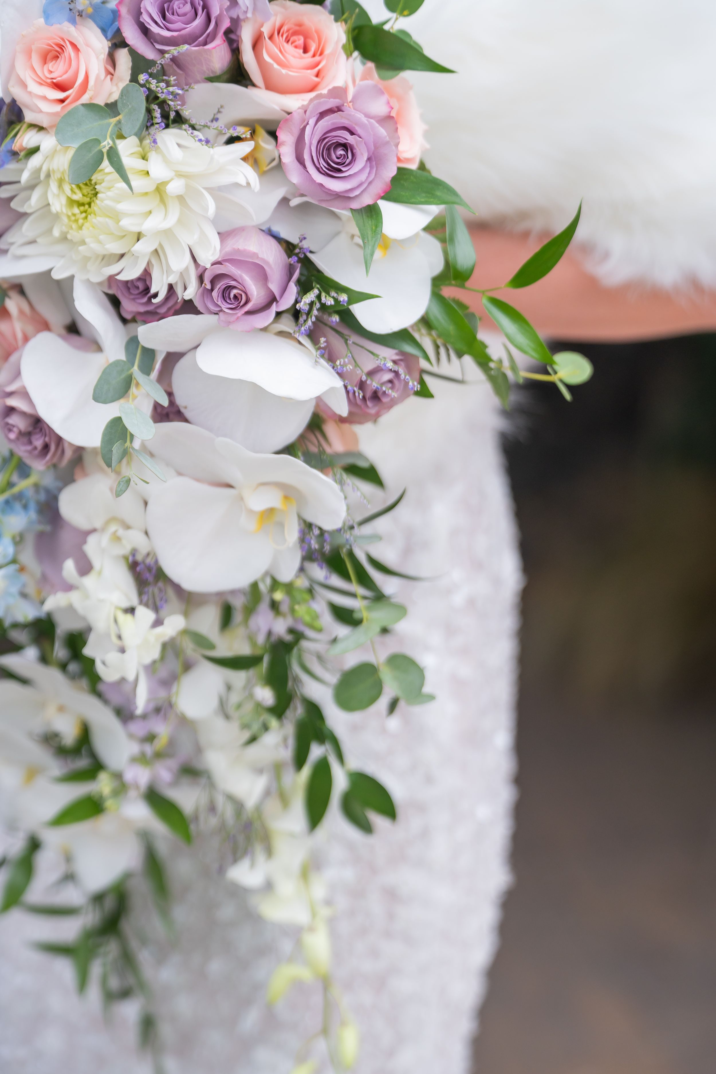 A romantic bouquet featuring white orchids, purple and pink roses with delicate eucalyptus leaves against a light background.