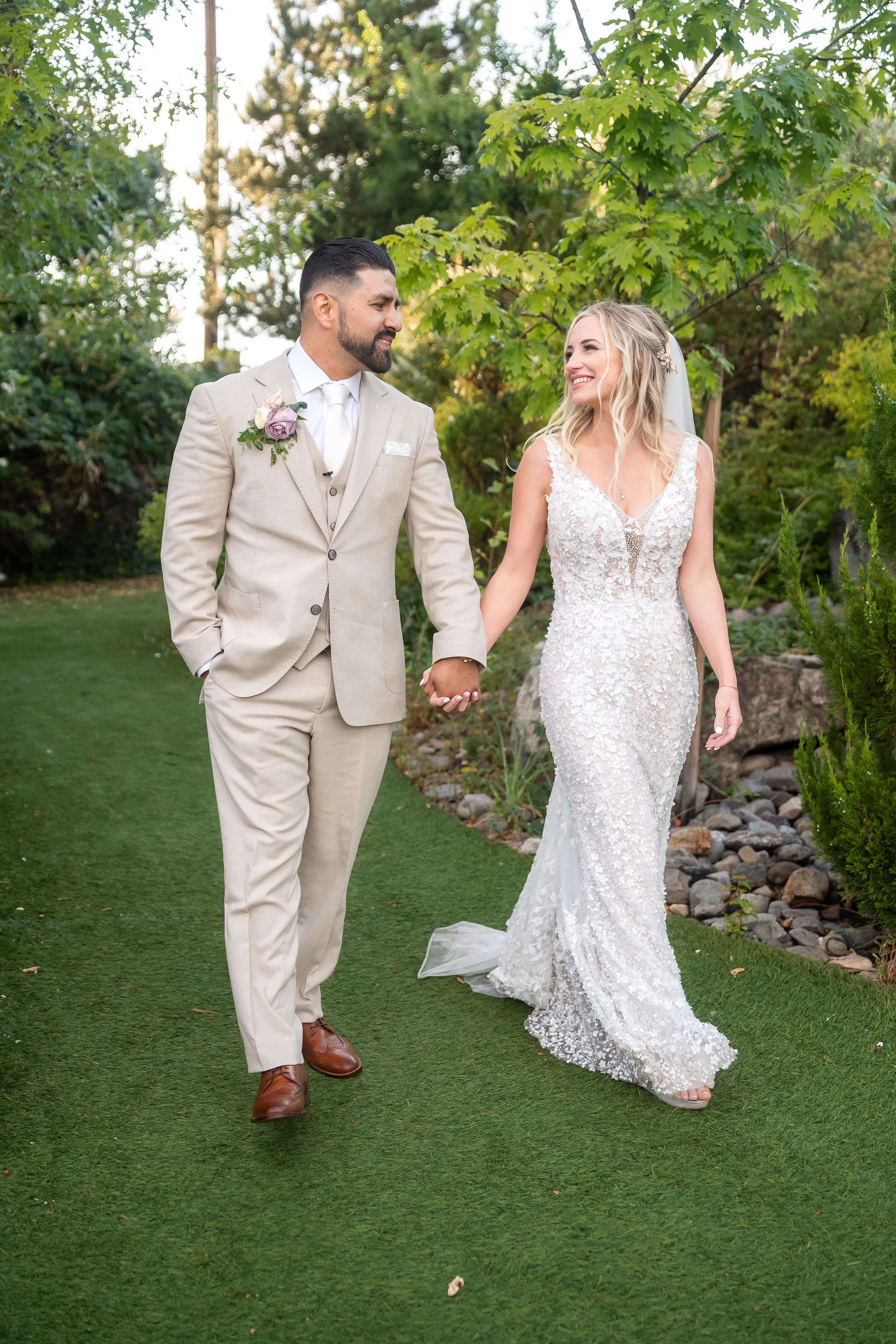 A bride and groom hold hands while walking on a garden path in elegant wedding attire.