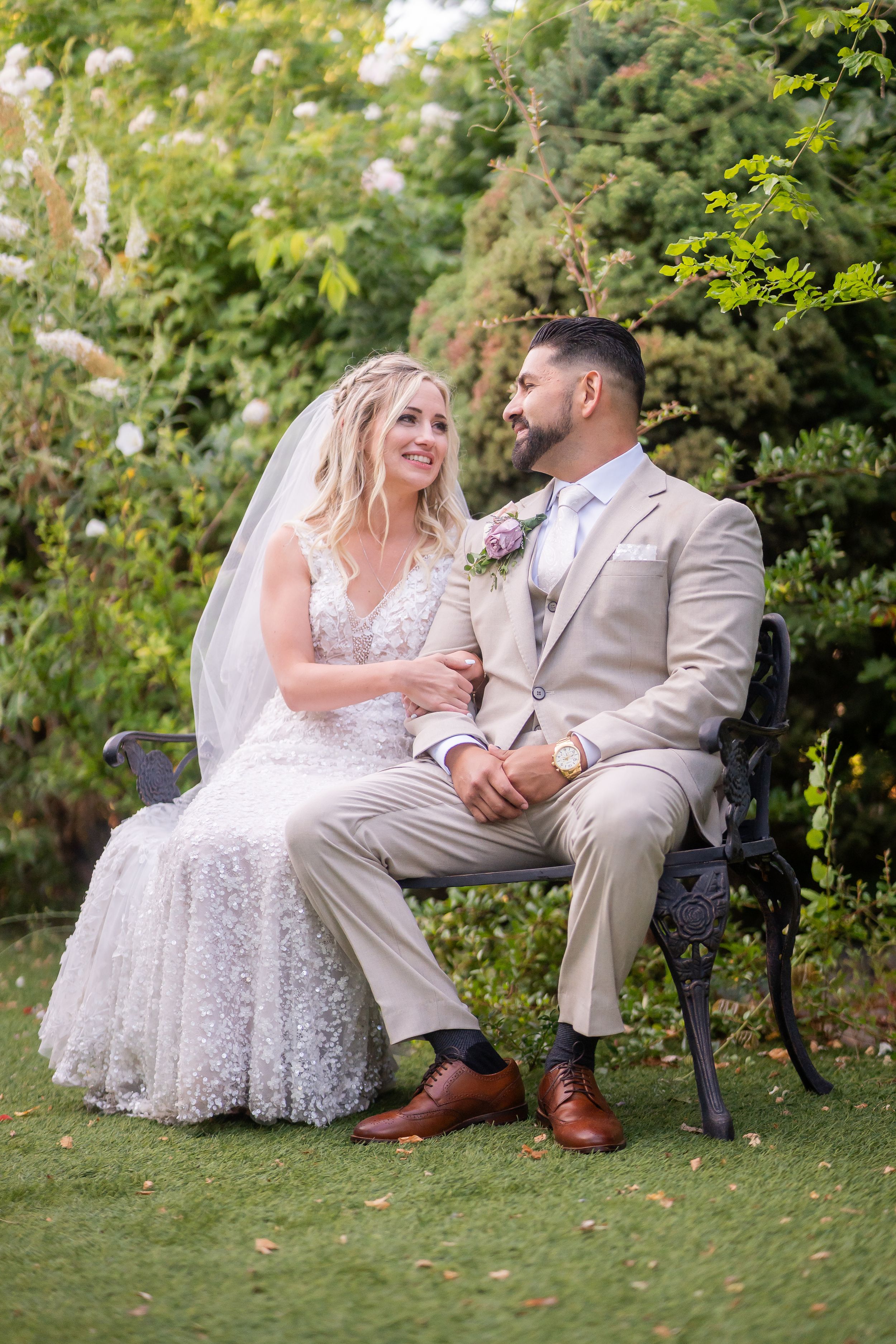 A couple in wedding attire sits together on a garden bench surrounded by greenery on their special day.