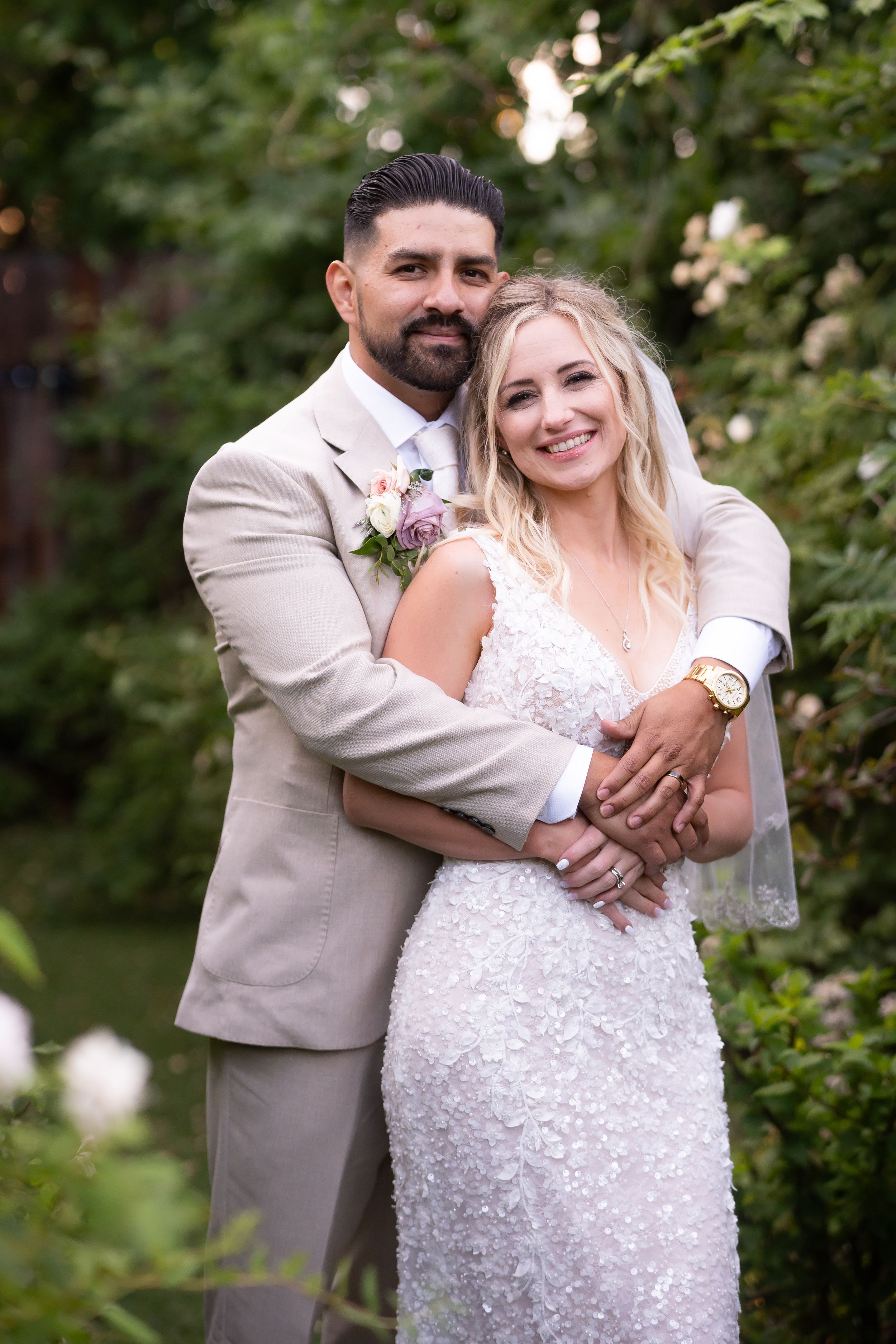 A beaming couple embraces in a garden setting, with the bride wearing a sparkly white gown and groom in a tan suit.