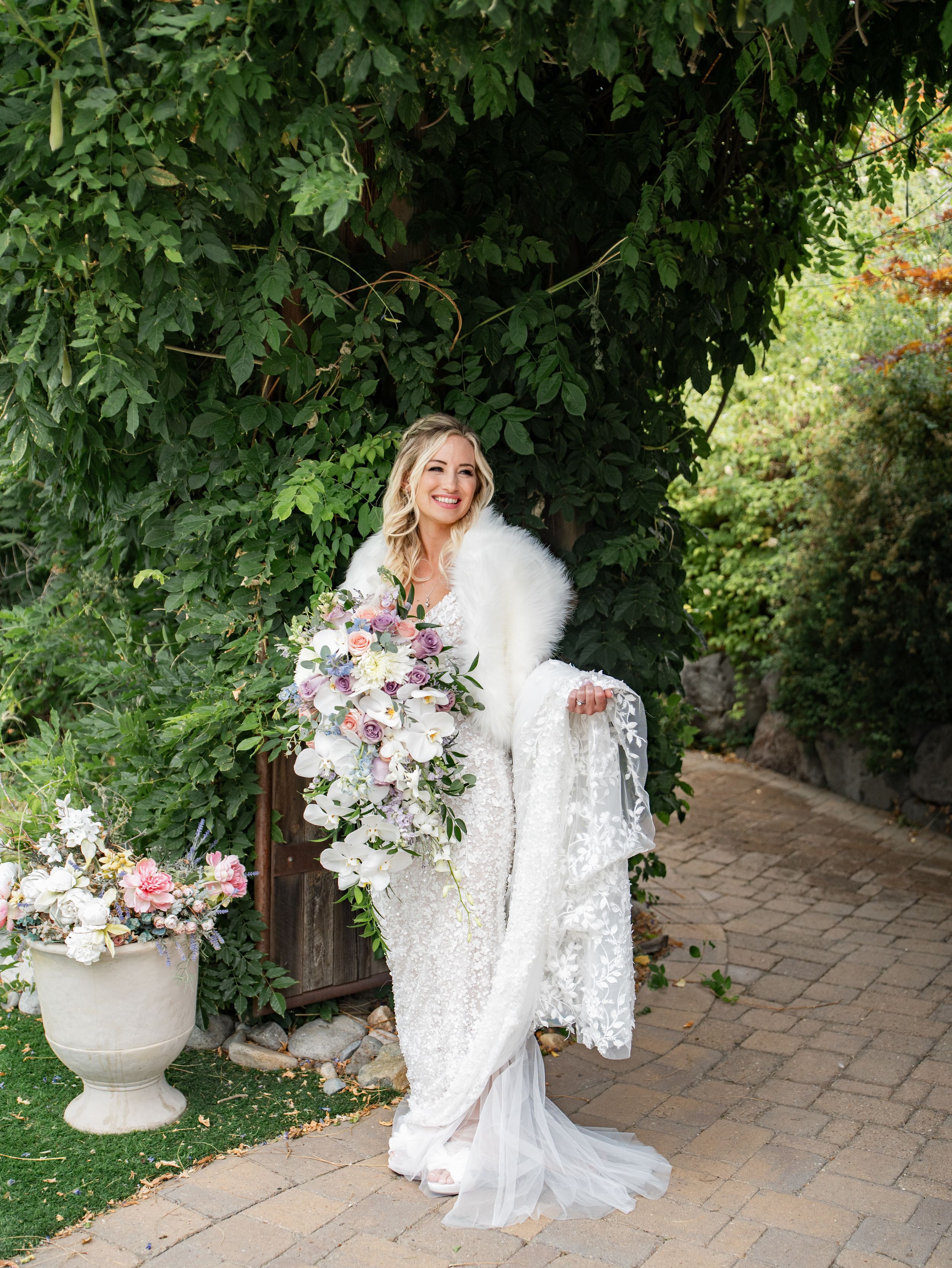 A bride in an elegant lace wedding gown and fur shawl holds a cascading purple and white bouquet in a garden setting at Las Cascadas, Reno.