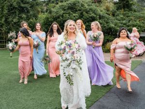 A bridal party in pastel dresses walks and laughs together on a lawn with lush greenery in the background.