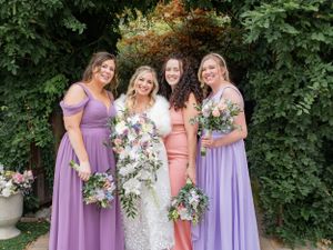 Bridesmaids in purple dresses and one in pink pose with a bride holding cascading floral bouquets in a garden setting at Las Cascadas
