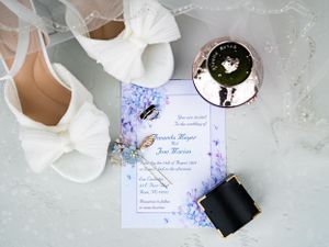 Wedding detail flatlay with white bridal shoes, floral invitation card, and black ring box on light gray surface.