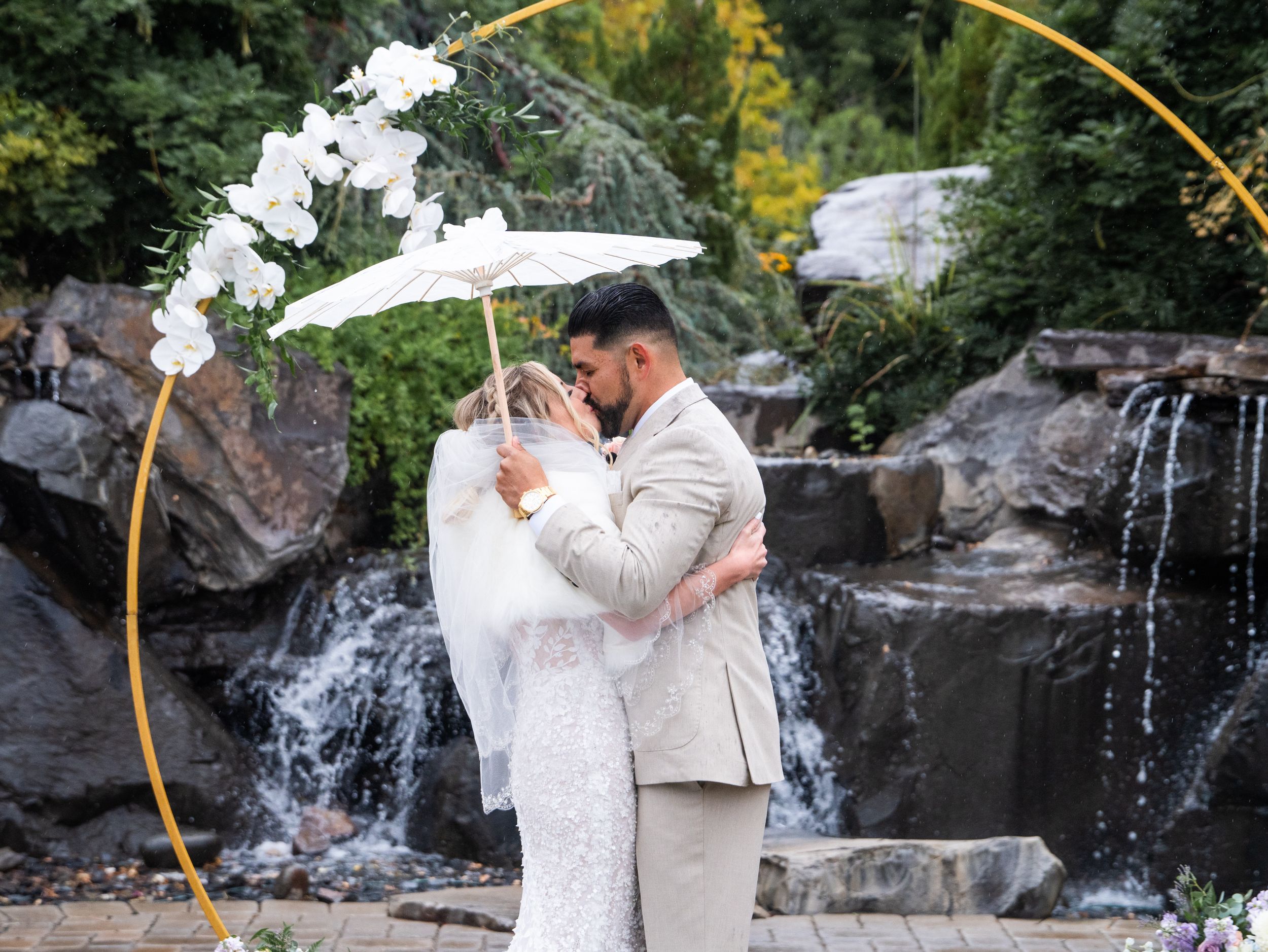 A couple shares a romantic moment under a floral arch with a cascading waterfall and natural rock backdrop in Reno at Las Cascadas.