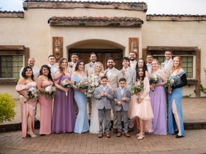 A wedding party in pastel colored dresses and suits poses together on a brick patio in front of a Spanish style building.