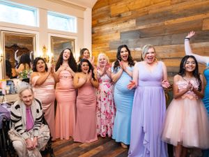 A group of bridesmaids in pastel-colored dresses smile and celebrate together against a rustic wooden wall at a bridal suite at Las Cascadas