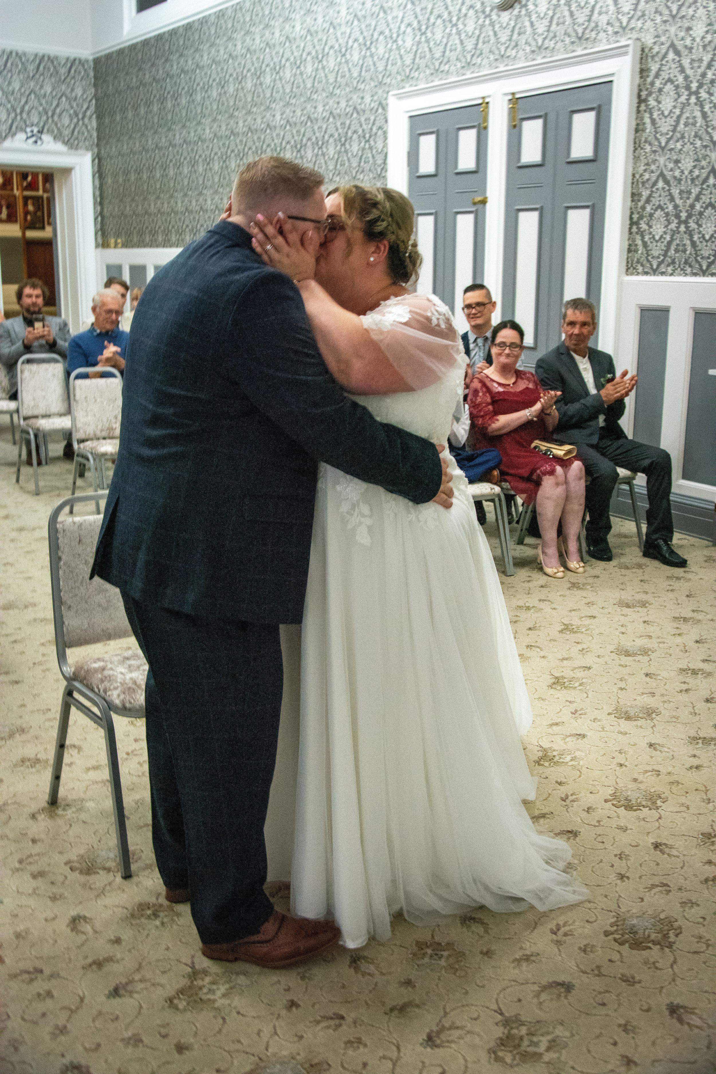 A passionate first kiss during a wedding ceremony with guests looking on in an elegant room.
