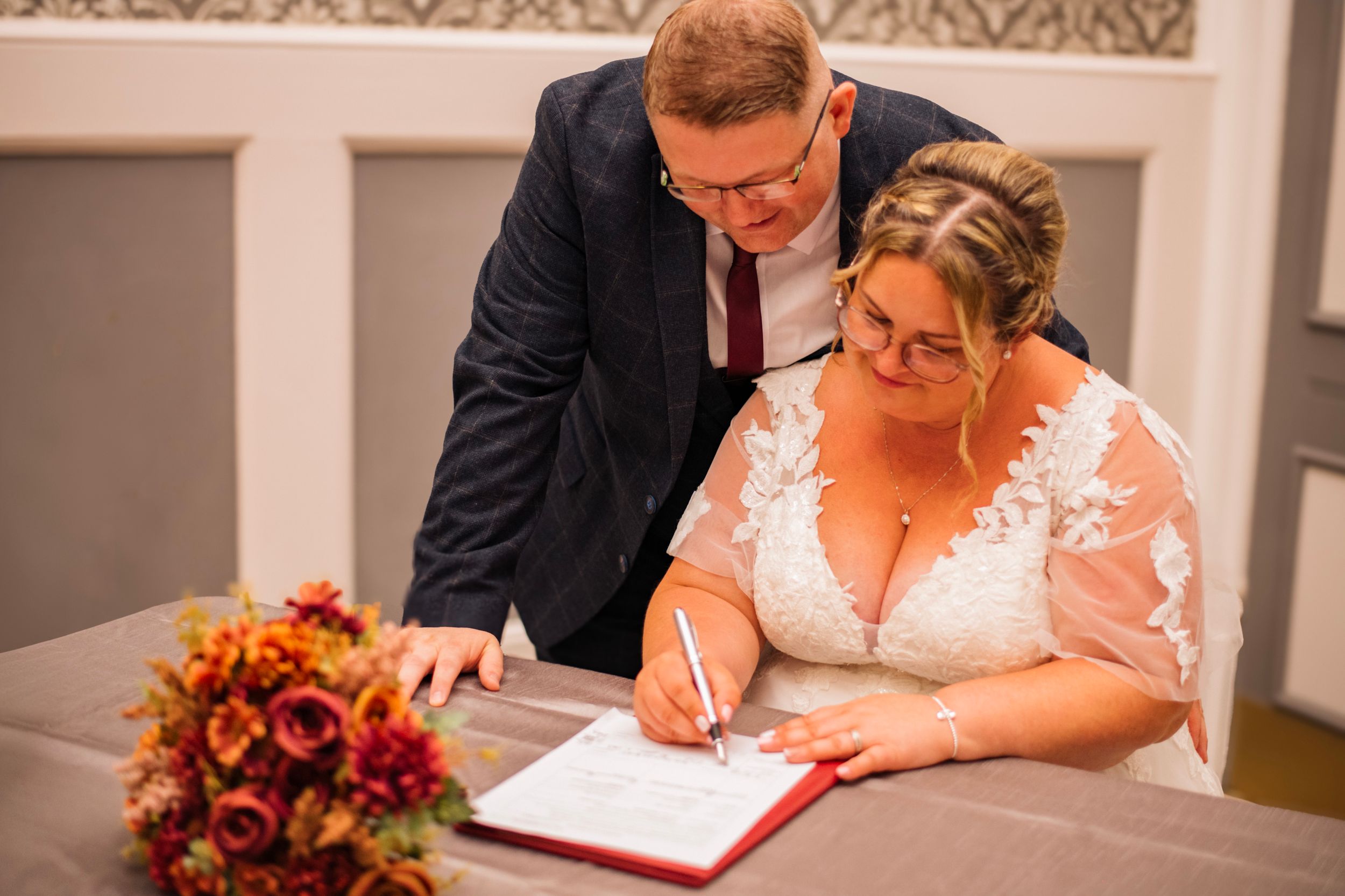 Wedding couple signing marriage documents at a table with autumn floral bouquet.