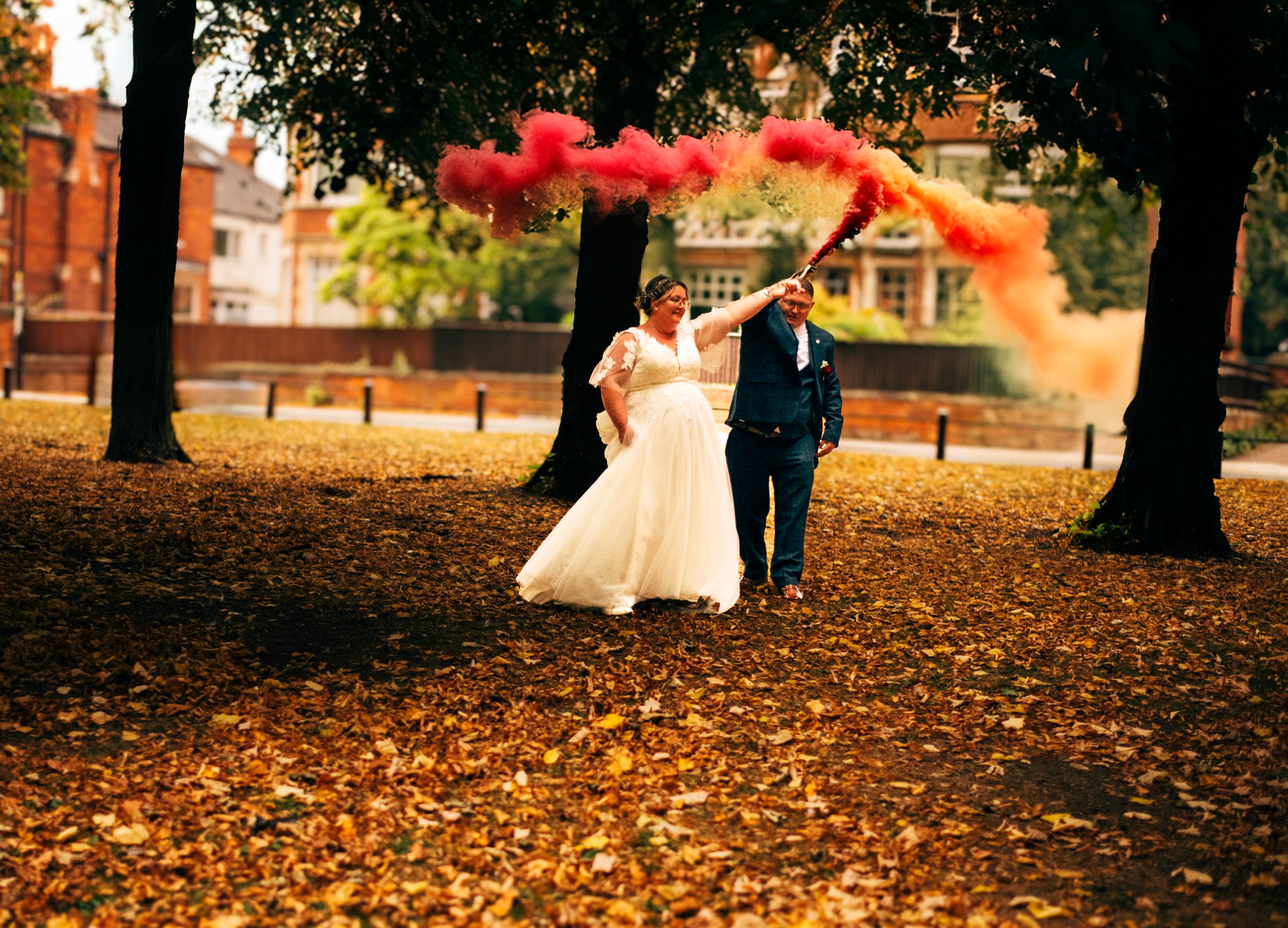 Newlyweds posing in autumn park with red smoke flares against fall foliage.