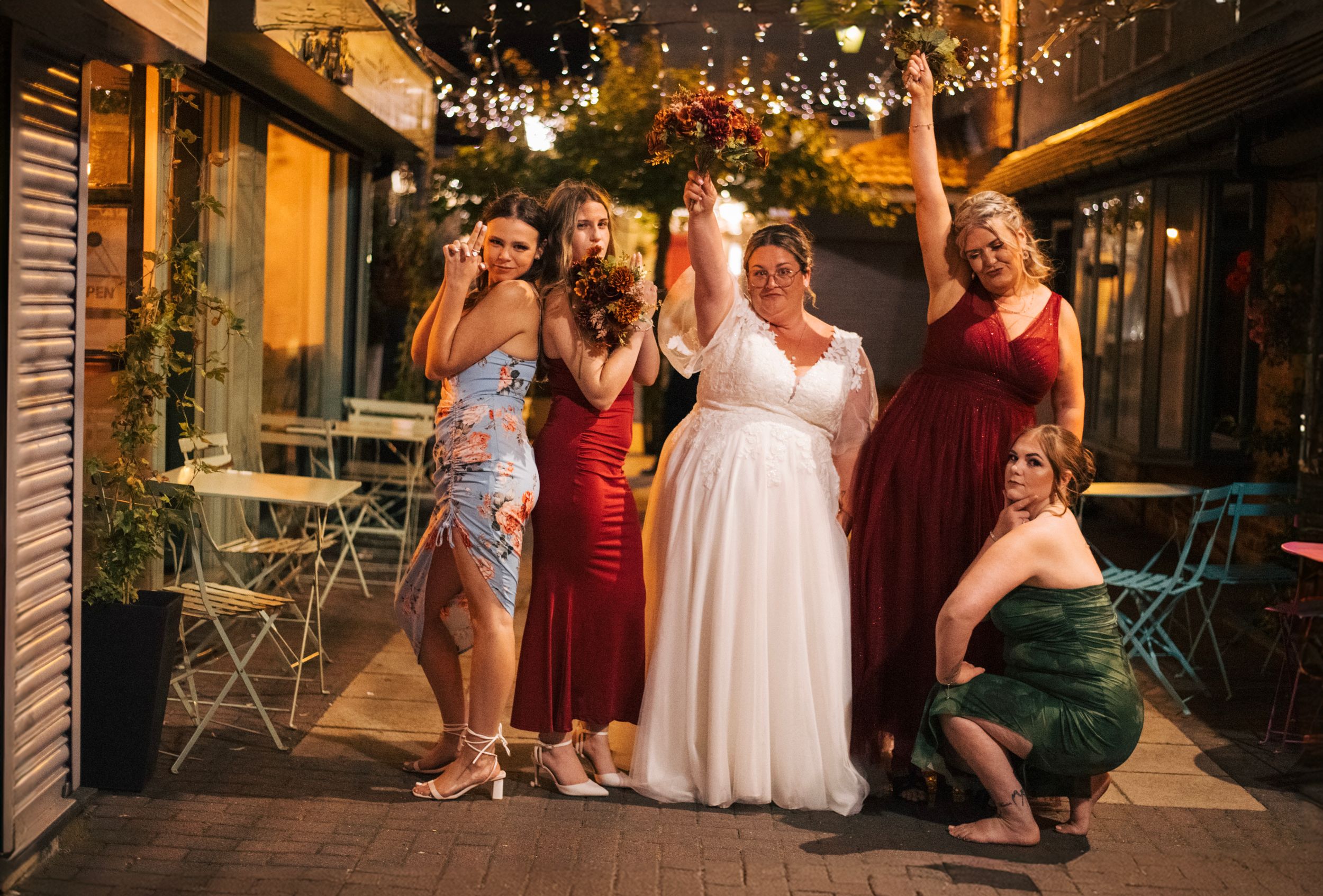 Bridal party posing together outdoors at sunset with string lights overhead.