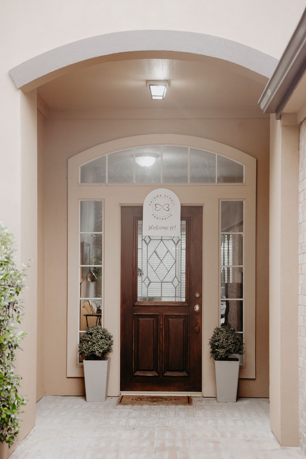 Elegant front entrance with dark wood door, decorative glass panels, and white planters under an arched beige portico.