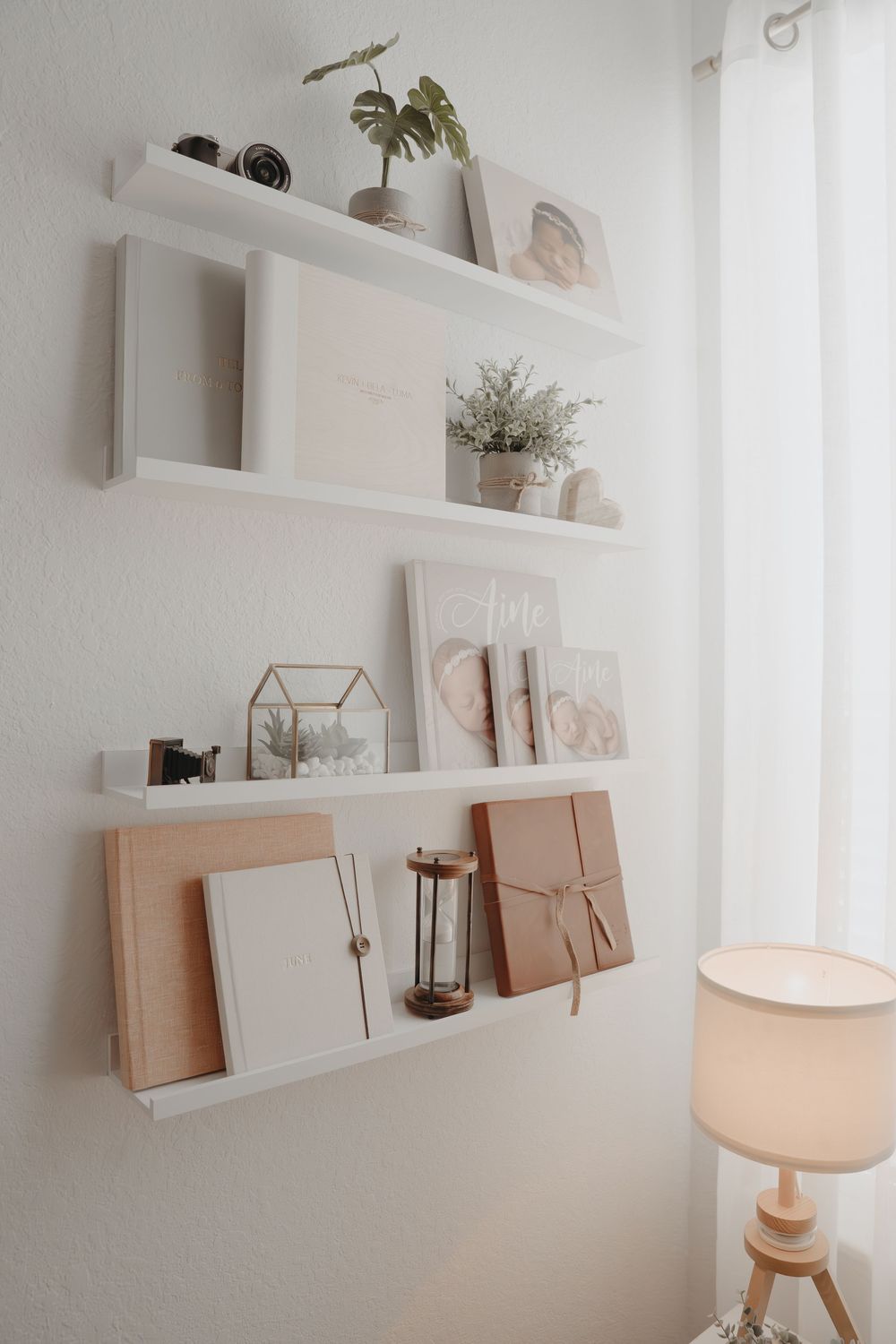 Minimalist white floating shelves displaying plants, decorative items, frames and artwork with a warm table lamp below.