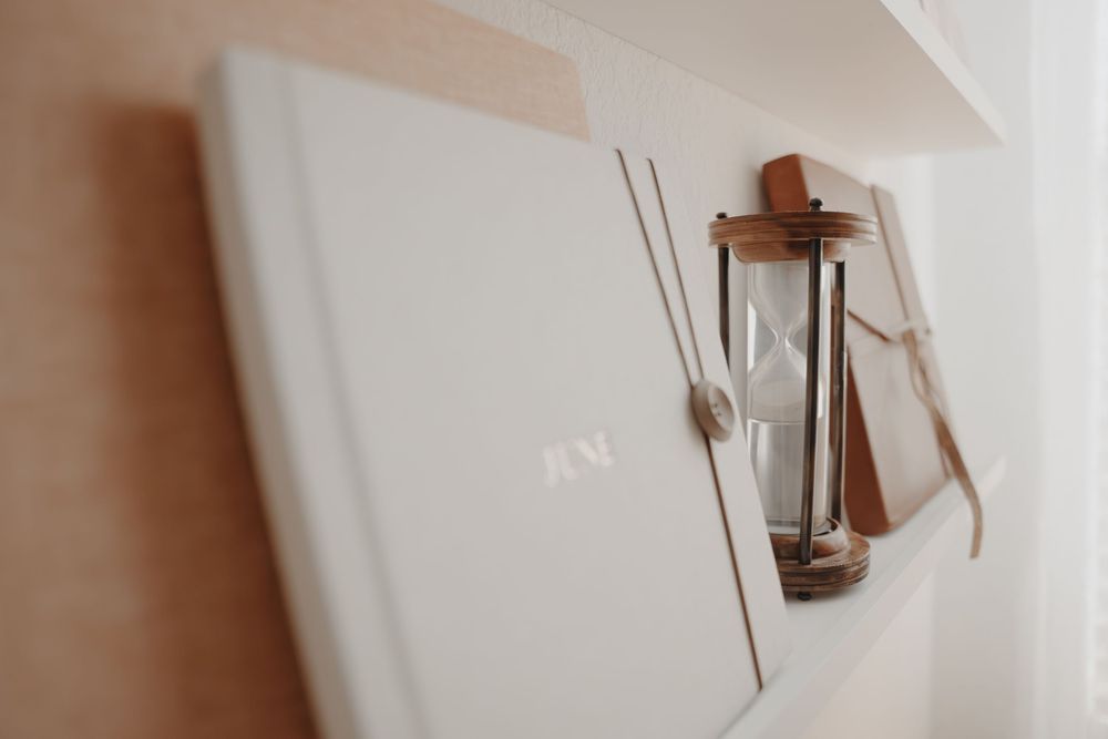 A glass and wood hourglass hangs on a white wall beside a book or notebook.