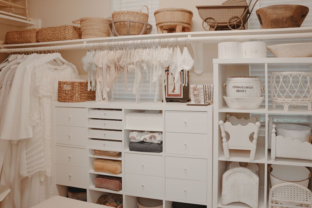 Organized white walk-in closet with woven baskets, drawers, and hanging clothes in a neutral color palette.