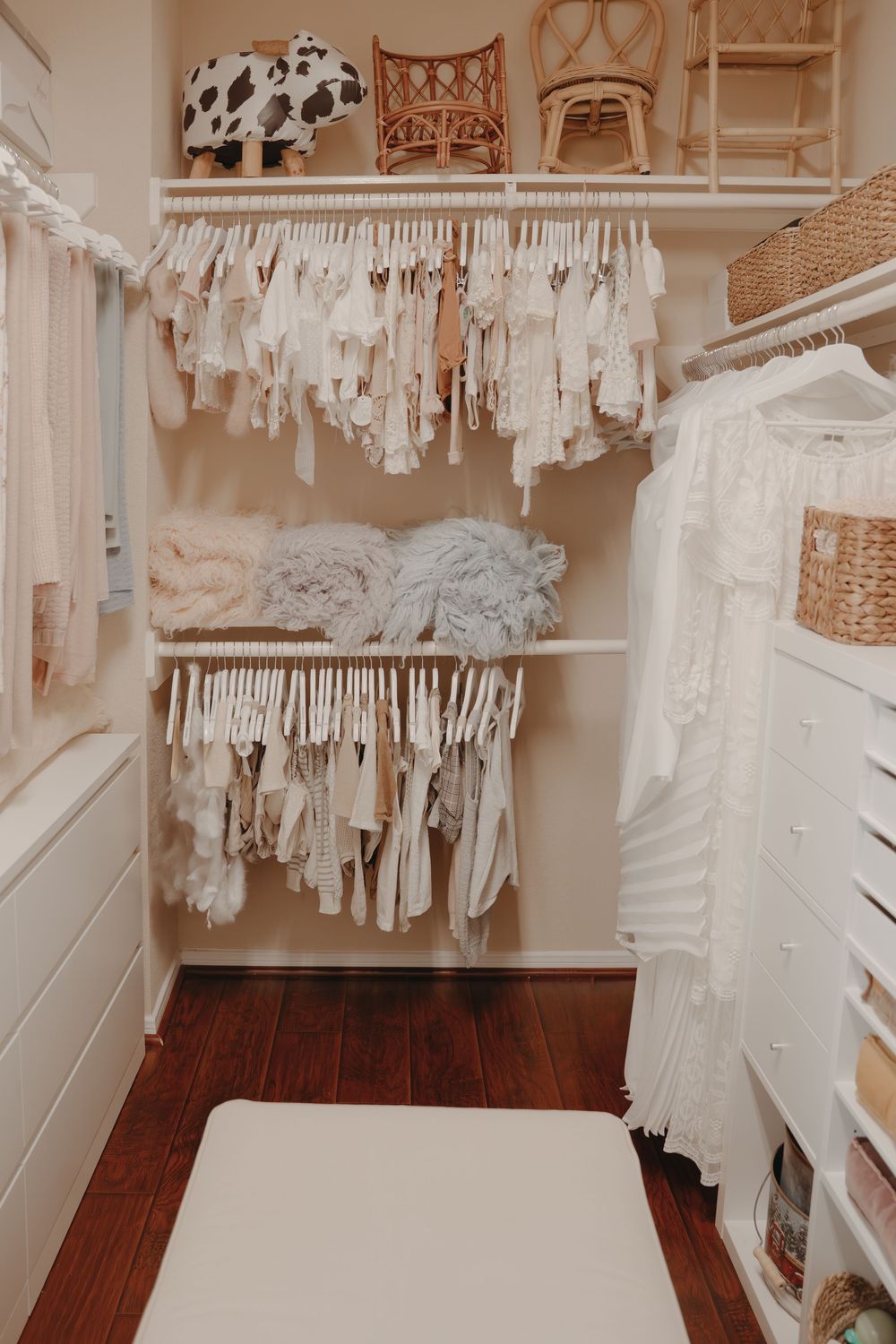 Neutral-toned nursery closet with organized white baby clothes hanging on rods and wicker baskets for storage.