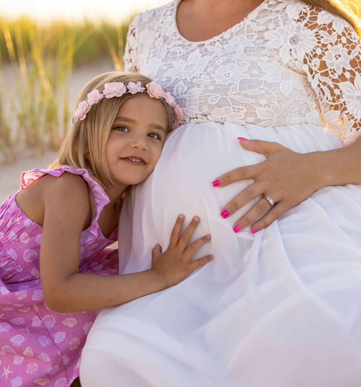 Young child in pink dress hugging pregnant woman's belly wearing white lace dress outdoors.