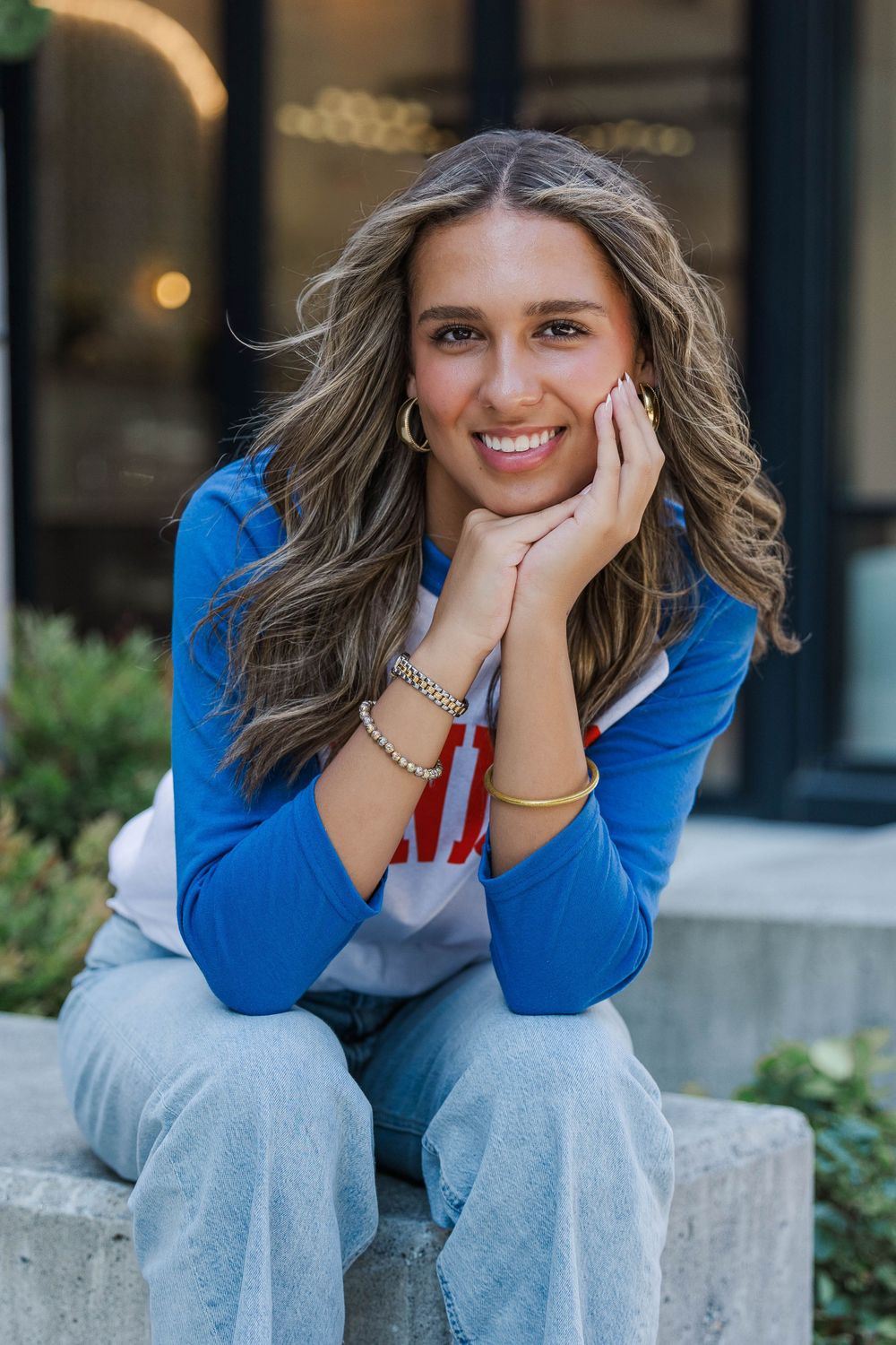 Smiling teenage girl with wavy hair poses on steps in blue SENIOR shirt and jeans for senior photos.