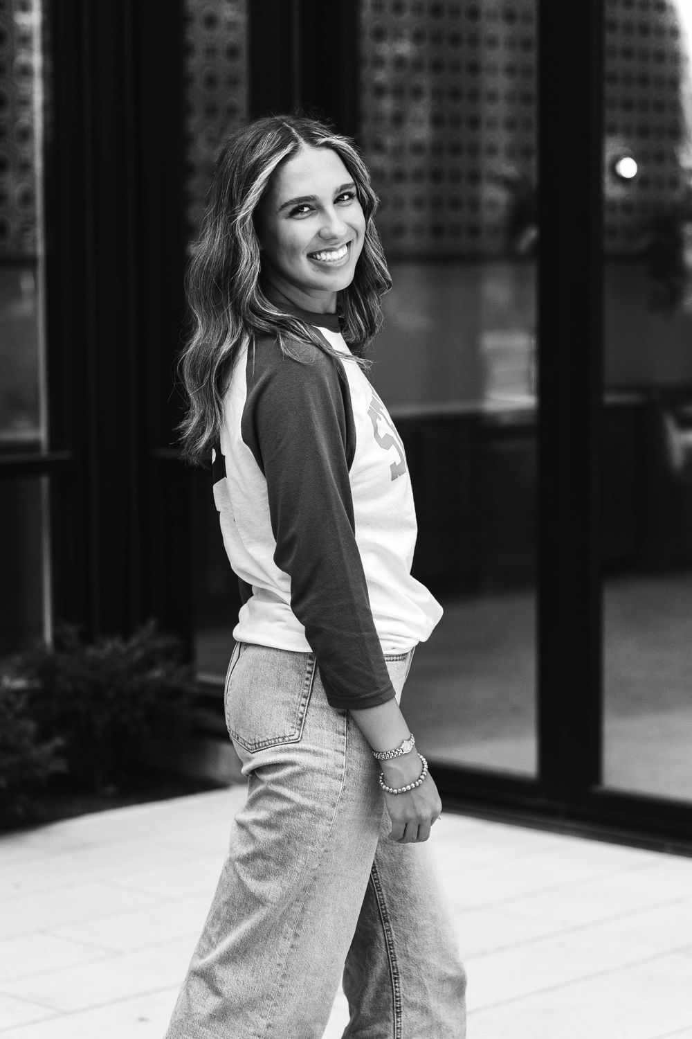Black and white senior photo of smiling teenage girl in baseball tee and wide-leg jeans outdoors.