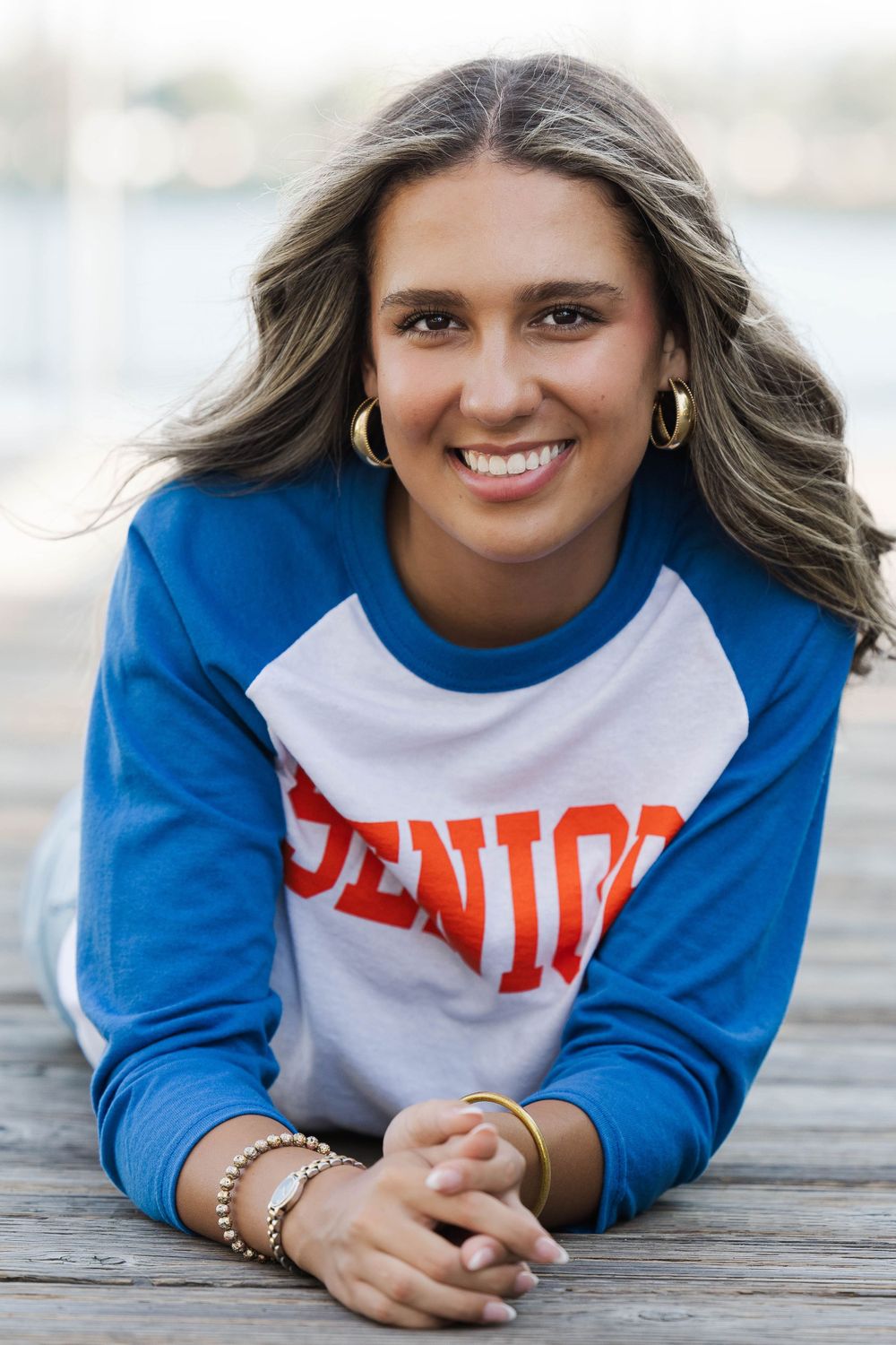 Smiling high school senior girl lying on wooden dock wearing blue and white SENIOR baseball shirt.