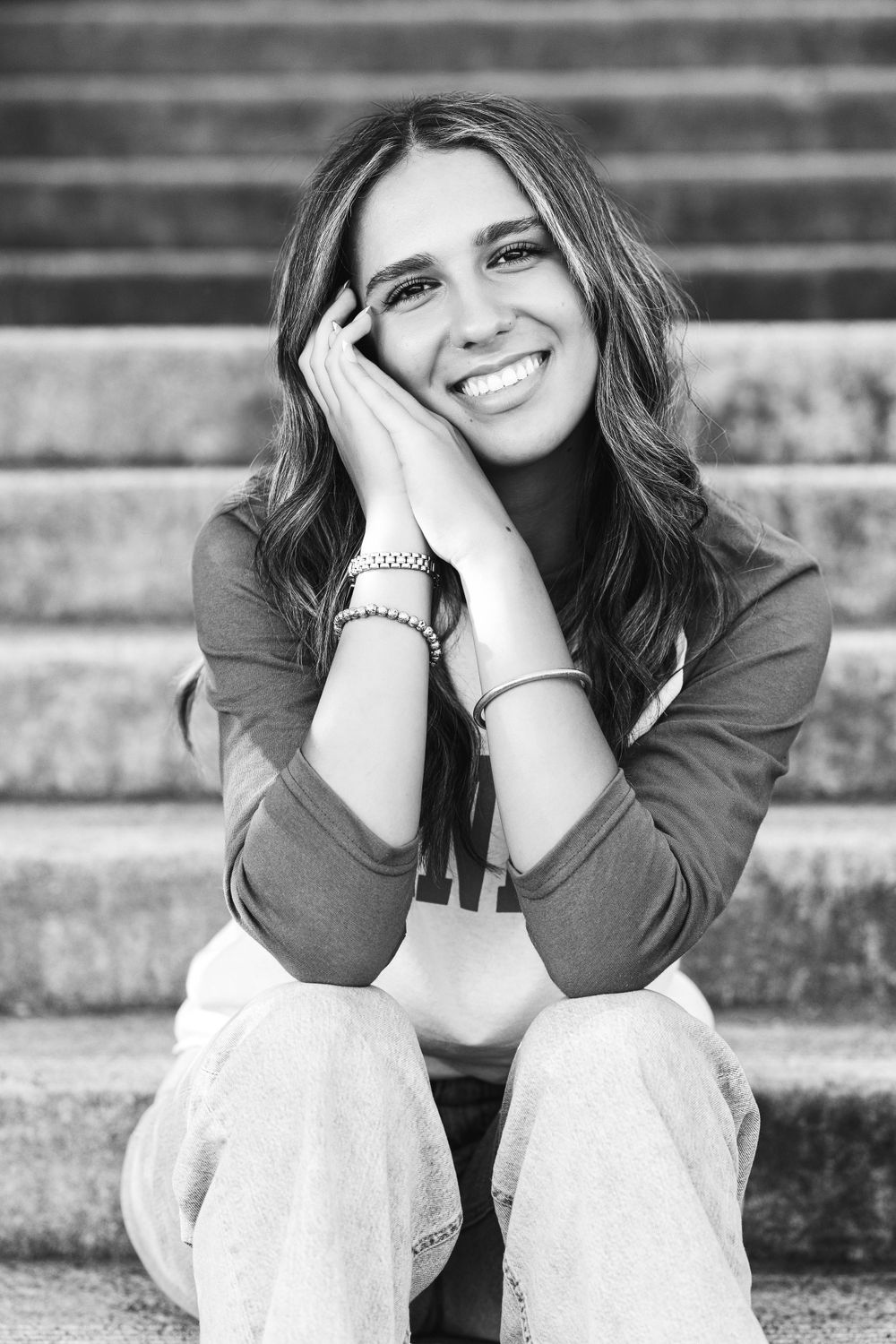 Black and white senior portrait of smiling teenage girl sitting on stone steps with hands on cheeks.