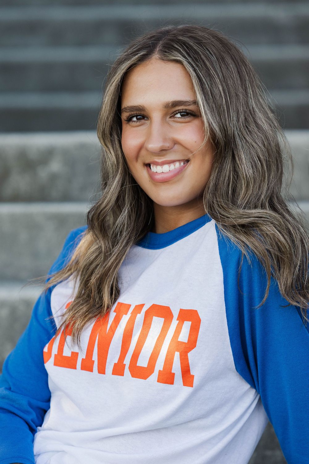 Close-up senior portrait of smiling teenage girl in blue and white SENIOR baseball tee on steps.