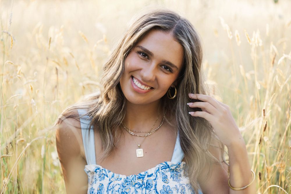 Smiling teenage girl in floral sundress sitting in golden wheat field for senior portrait session.