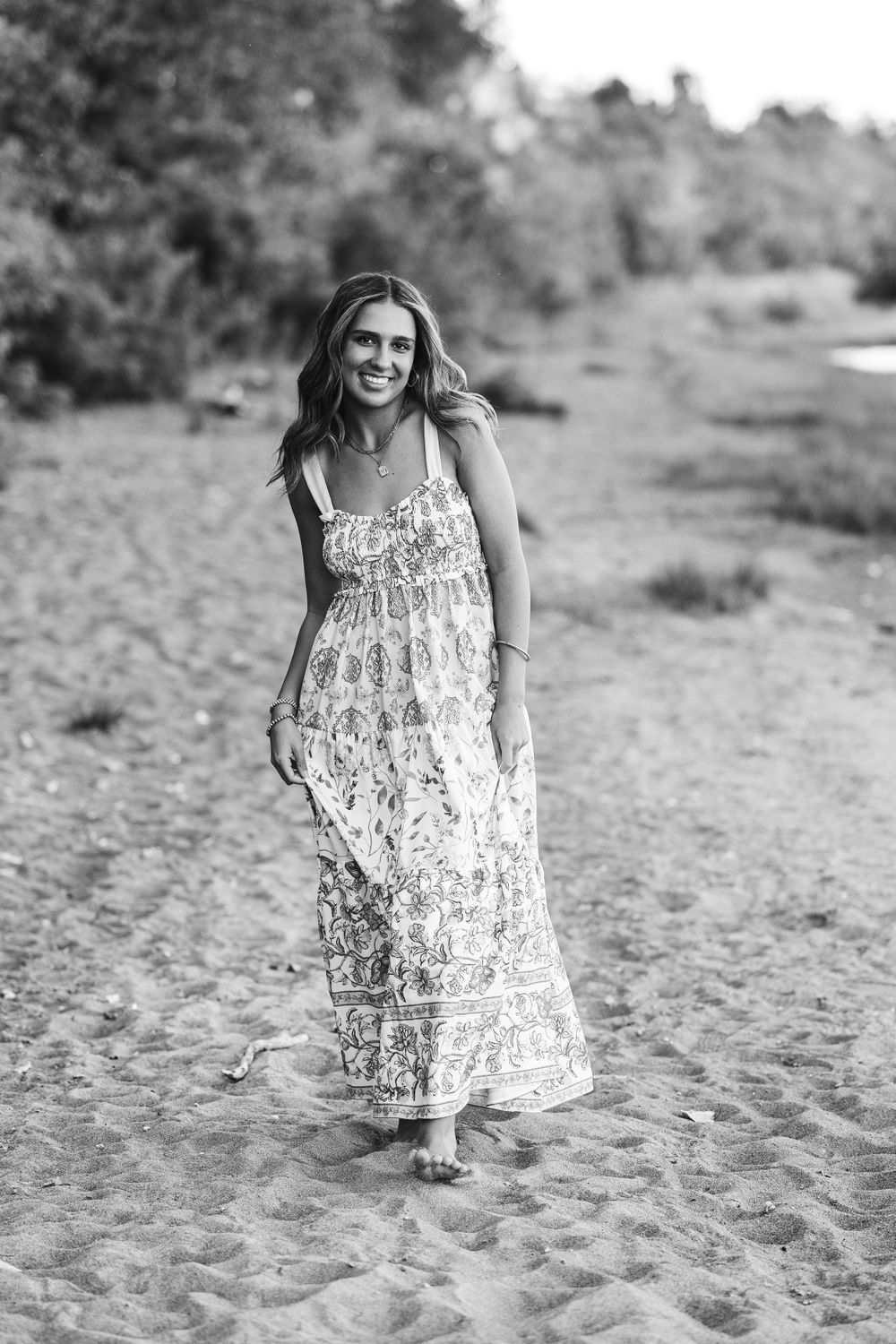 Black and white senior photo of smiling teenage girl in maxi dress walking barefoot on sandy beach.