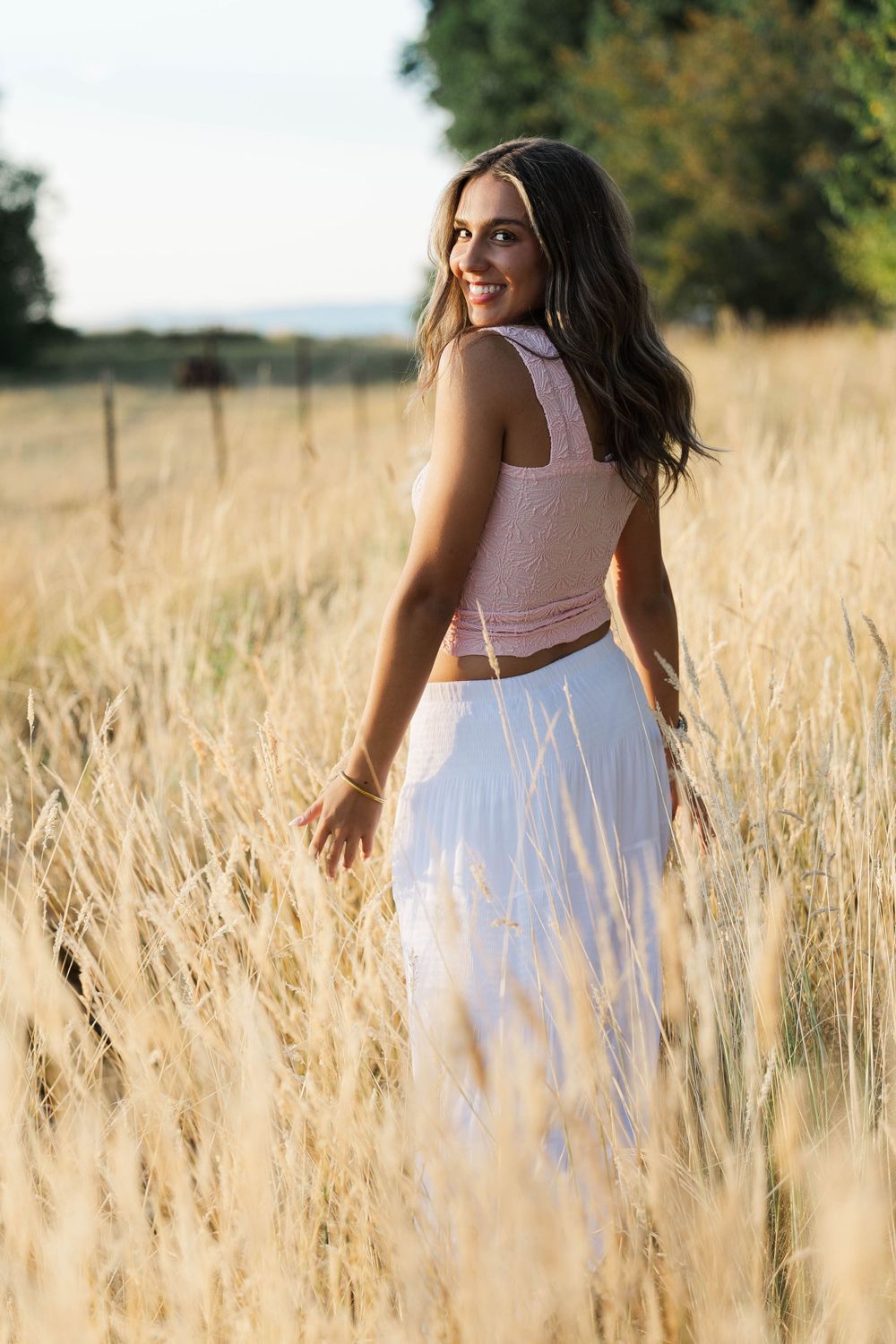Smiling teenage girl in pink crop top and white skirt walking through golden field for senior photos.