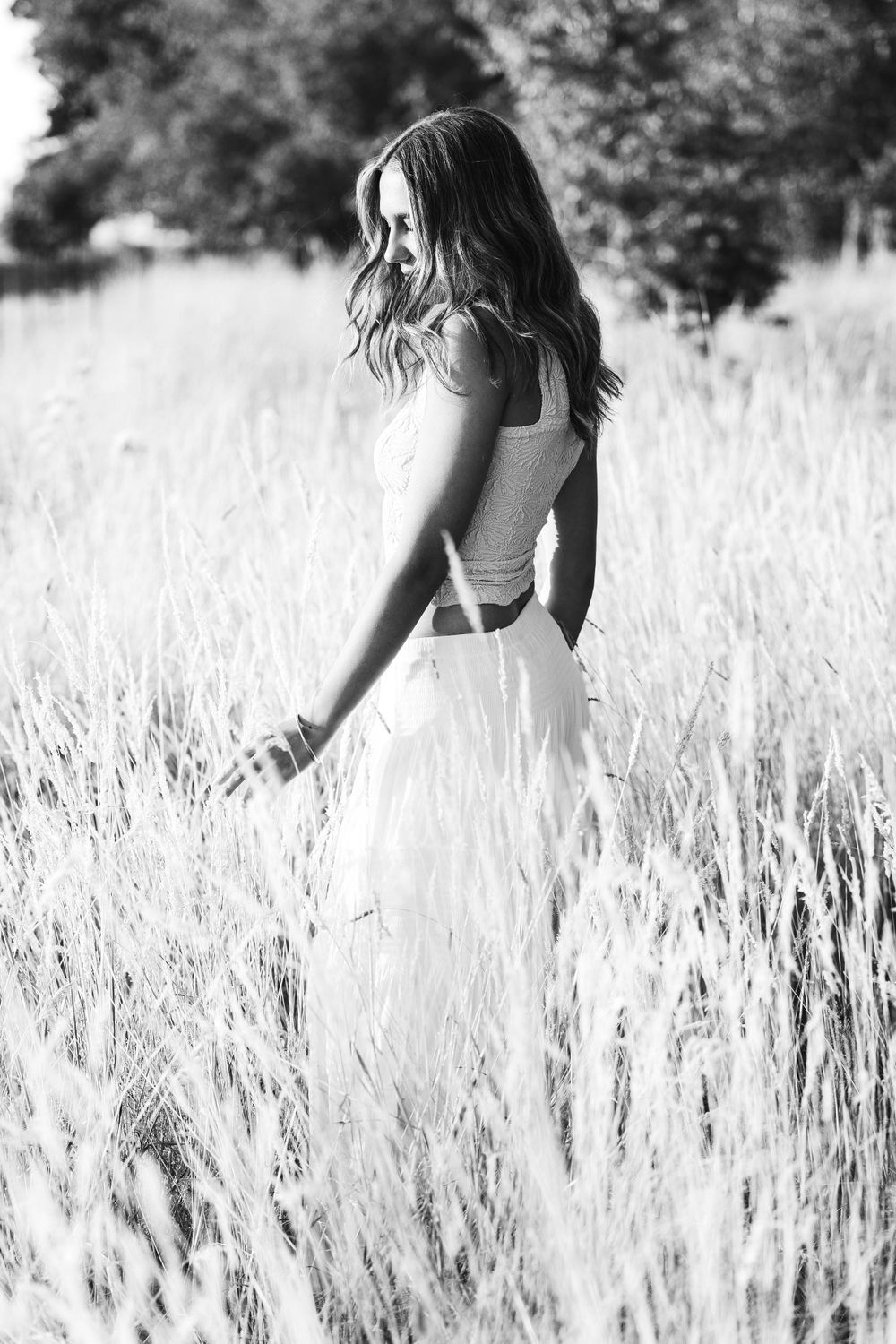 Young woman in white dress walking through tall golden grass field in dreamy black and white portrait.