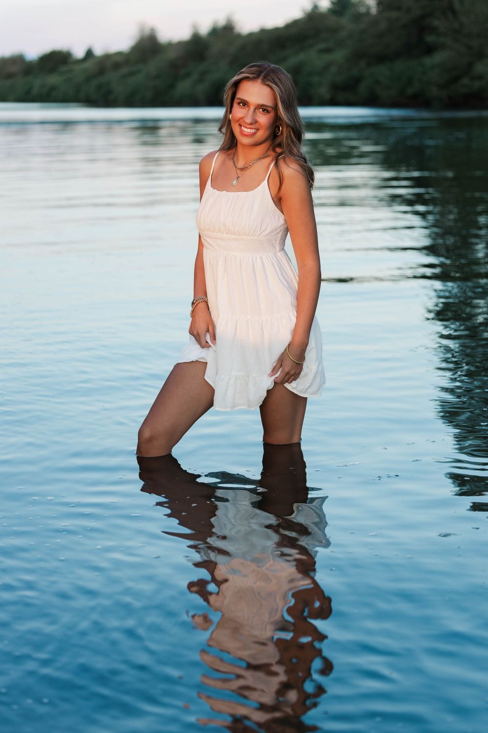 Smiling young woman in white sundress standing in calm lake water at sunset.