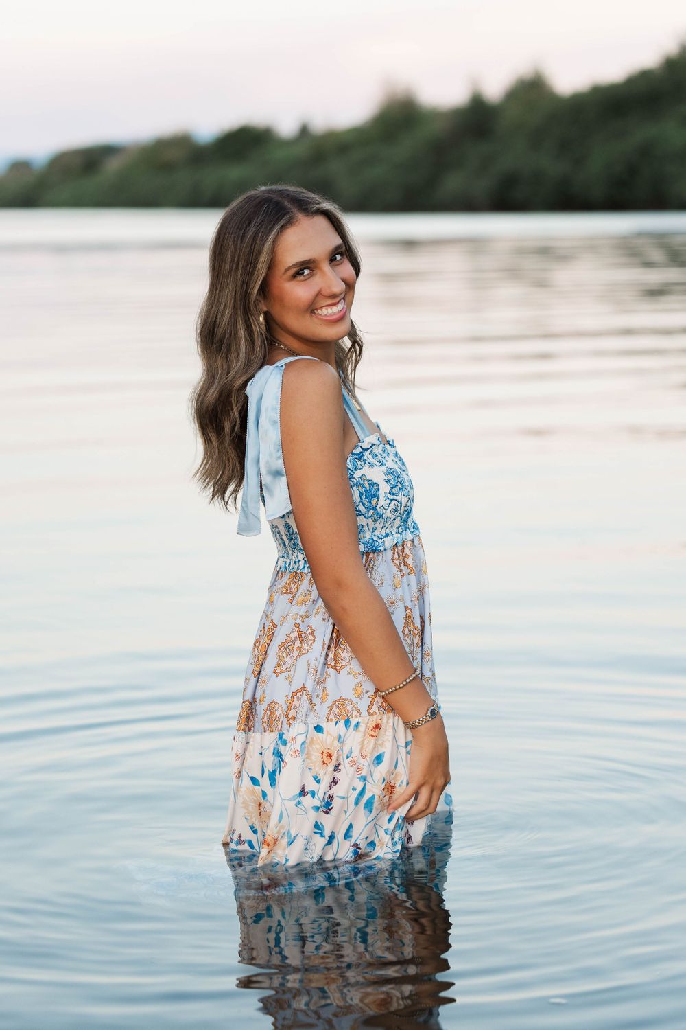 Happy young woman in floral boho dress standing in lake water smiling over shoulder at sunset.