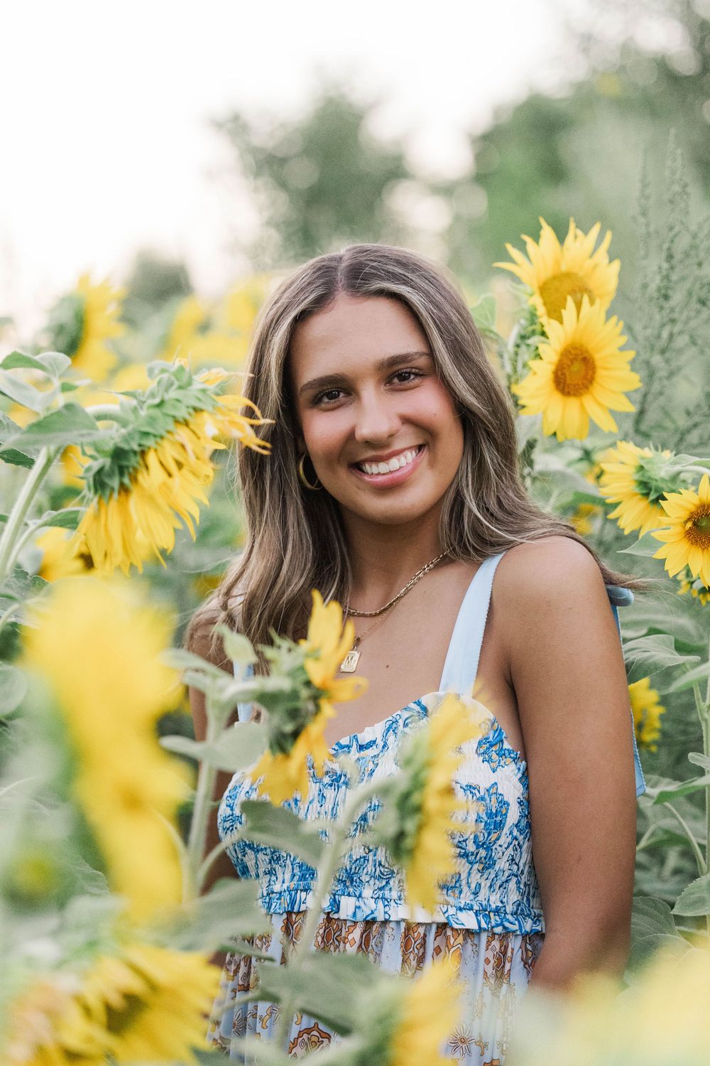 Smiling teenage girl in blue floral dress posing among bright yellow sunflowers in a garden.
