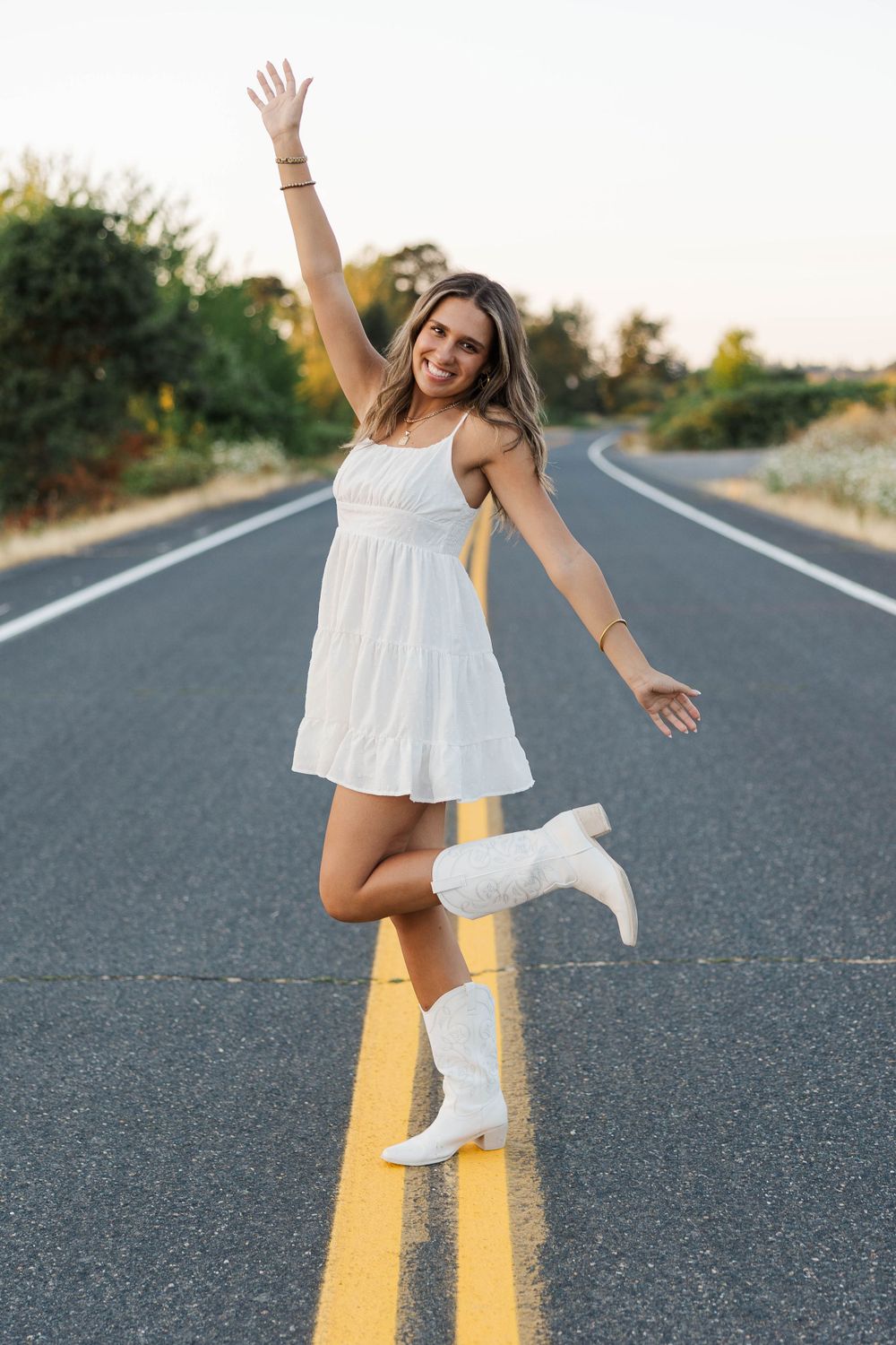 Joyful young woman in white mini dress and cowboy boots posing on open road at sunset.
