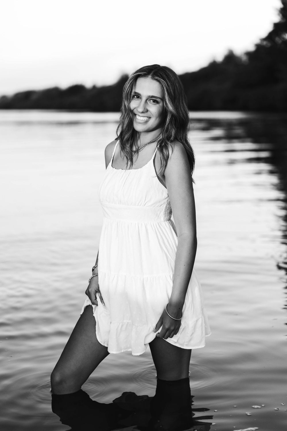 Smiling young woman in white sundress standing in lake water in black and white portrait.