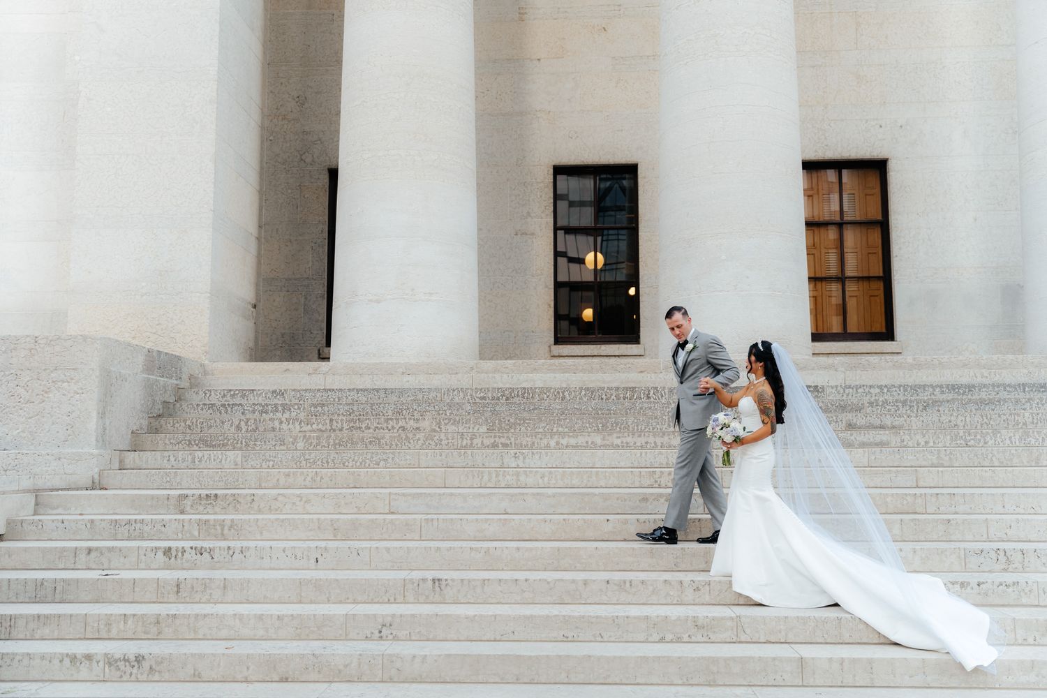 A couple in wedding attire poses on grand marble steps with white columns in the background.