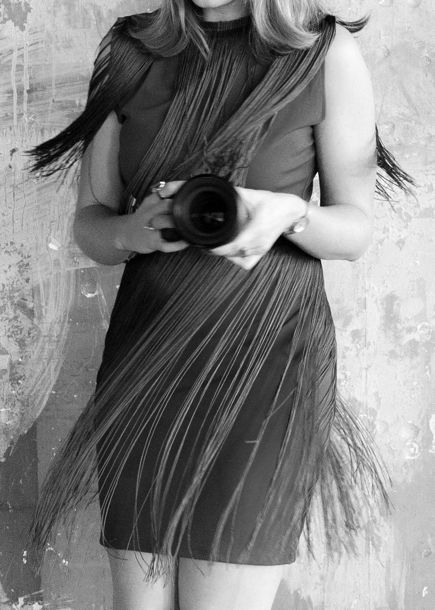 Black and white portrait of a fringe dress detail with camera against textured wall.