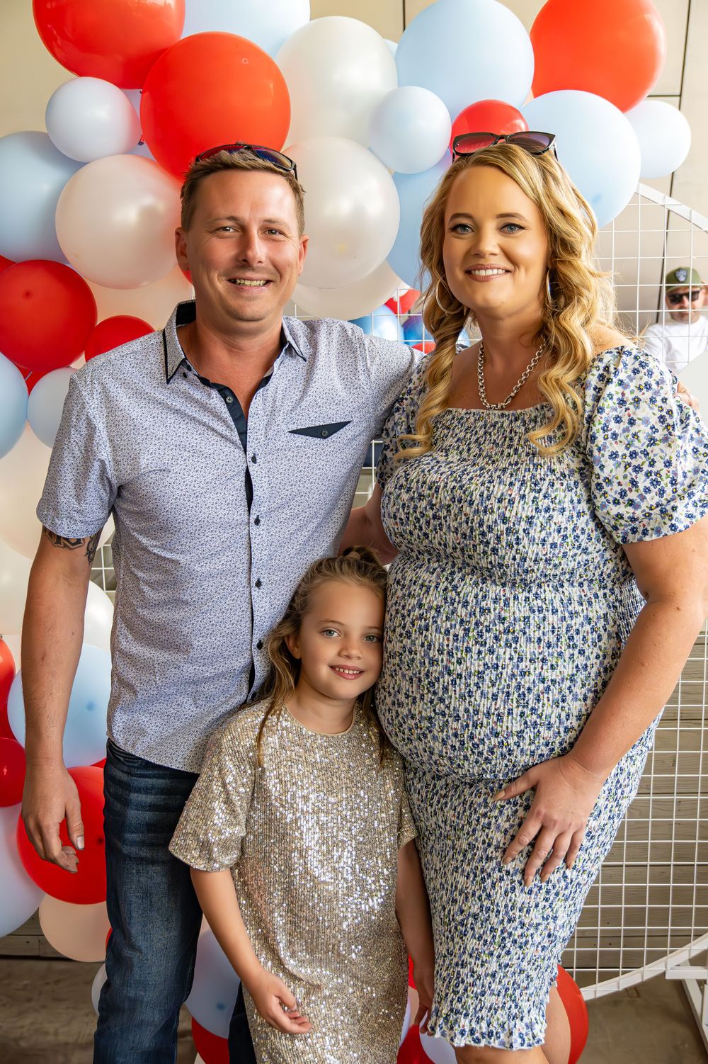Family of three posing in front of festive balloon backdrop at celebration.