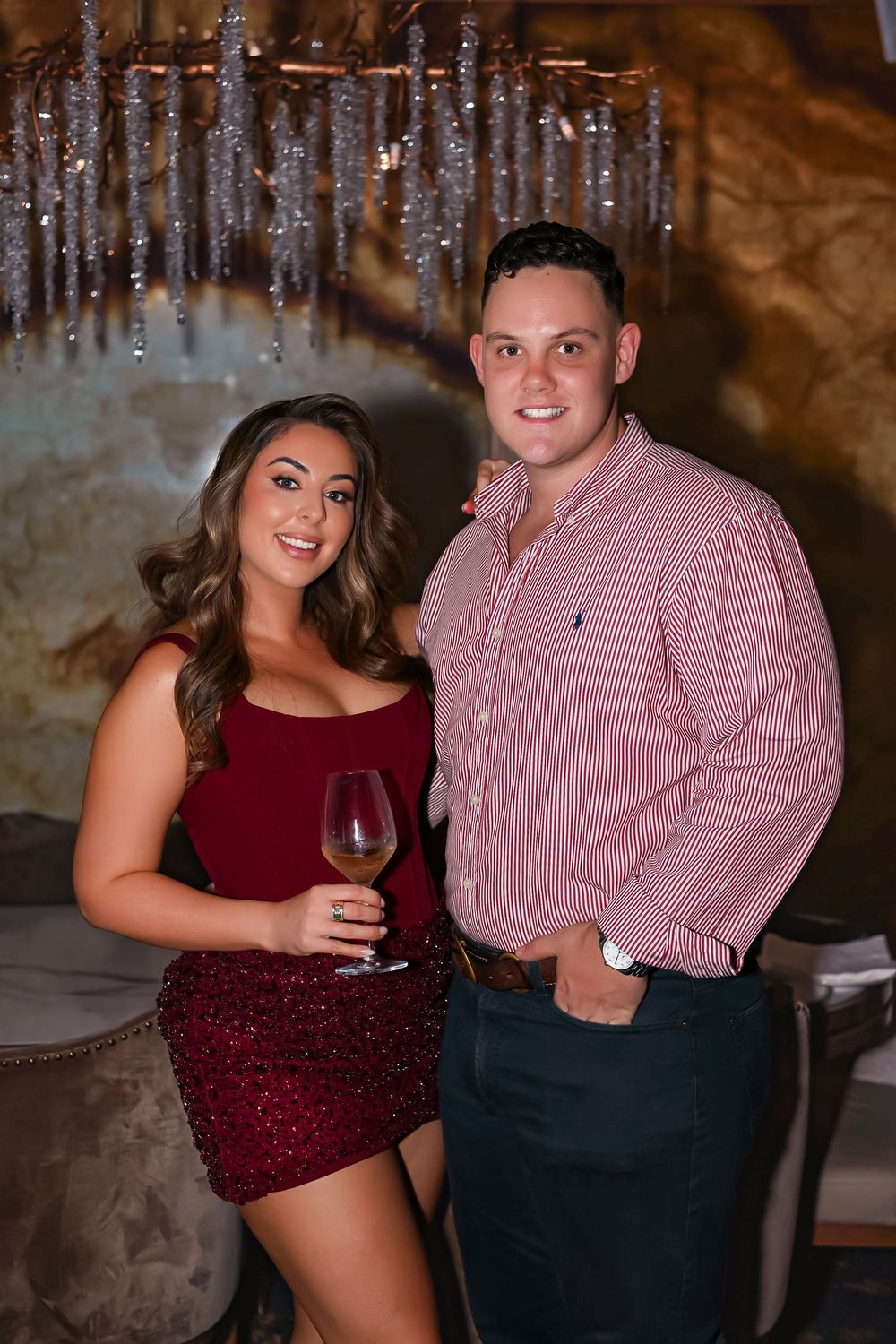 A couple poses elegantly at a restaurant, woman in red sequin dress holding wine, man in pink striped shirt smiling.