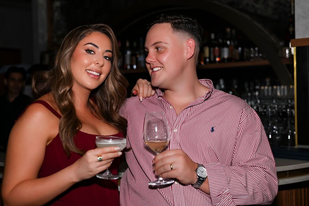 Couple smiling and holding wine glasses at a bar, woman in red dress, man in pink striped shirt.