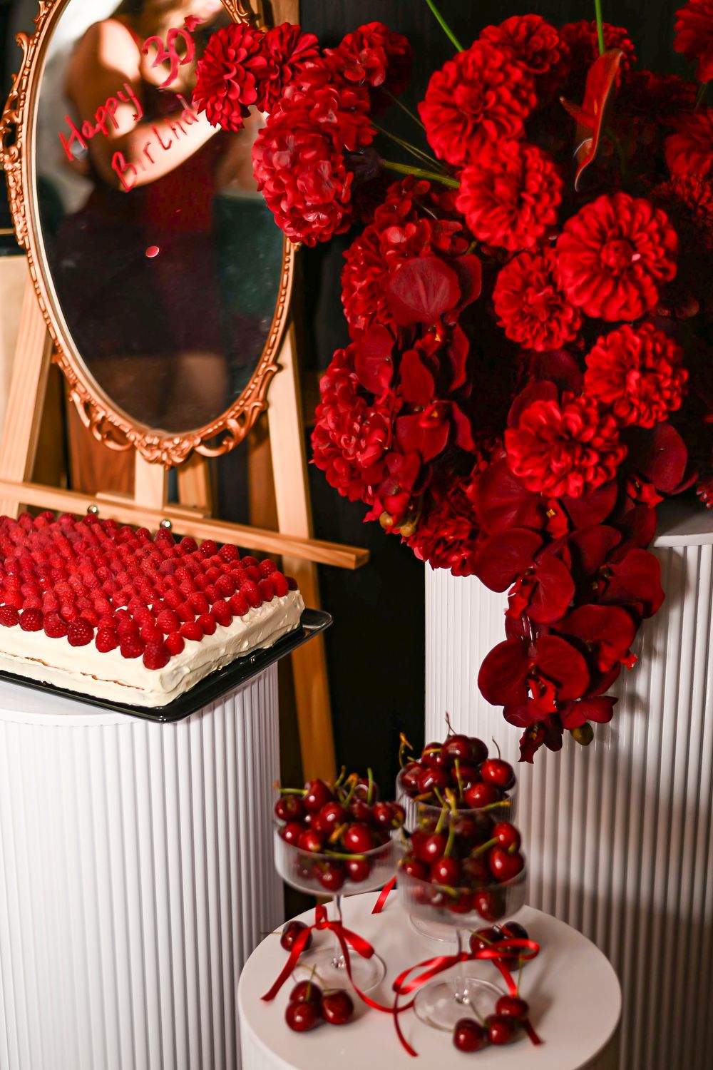 Elegant red floral arrangement with cherries, raspberry cake, and ornate mirror on white pedestals.