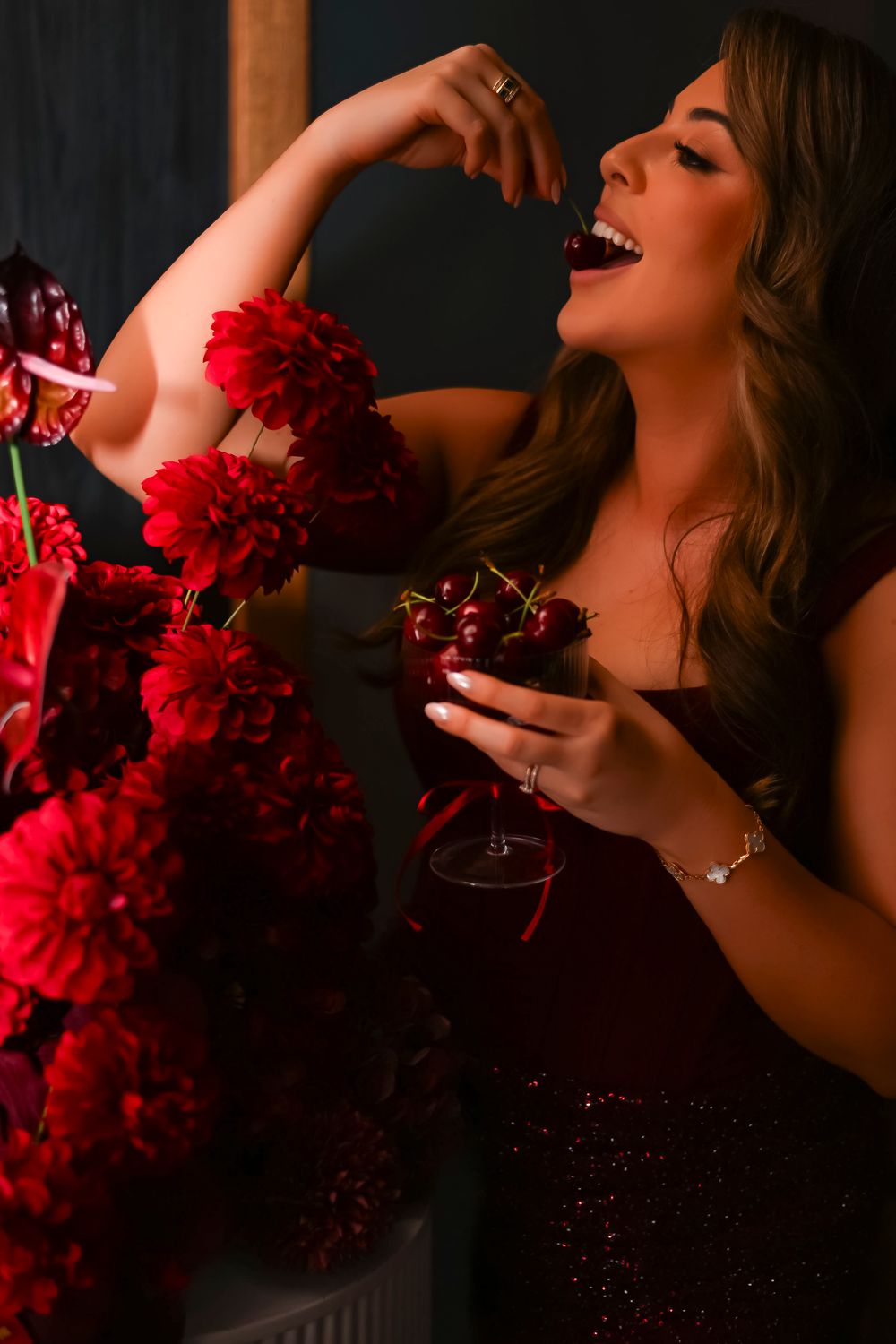A smiling woman in a red sequin dress holds cherries near red dahlia flowers in a dark, moody setting.